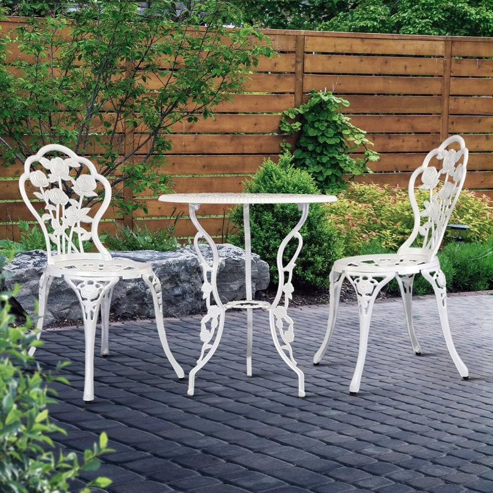 White aluminium bistro table and chairs set placed on stone patio surrounded by greenery.