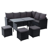 Outdoor dining sofa set with black wicker and beige cushions arranged on patio beside green plants.