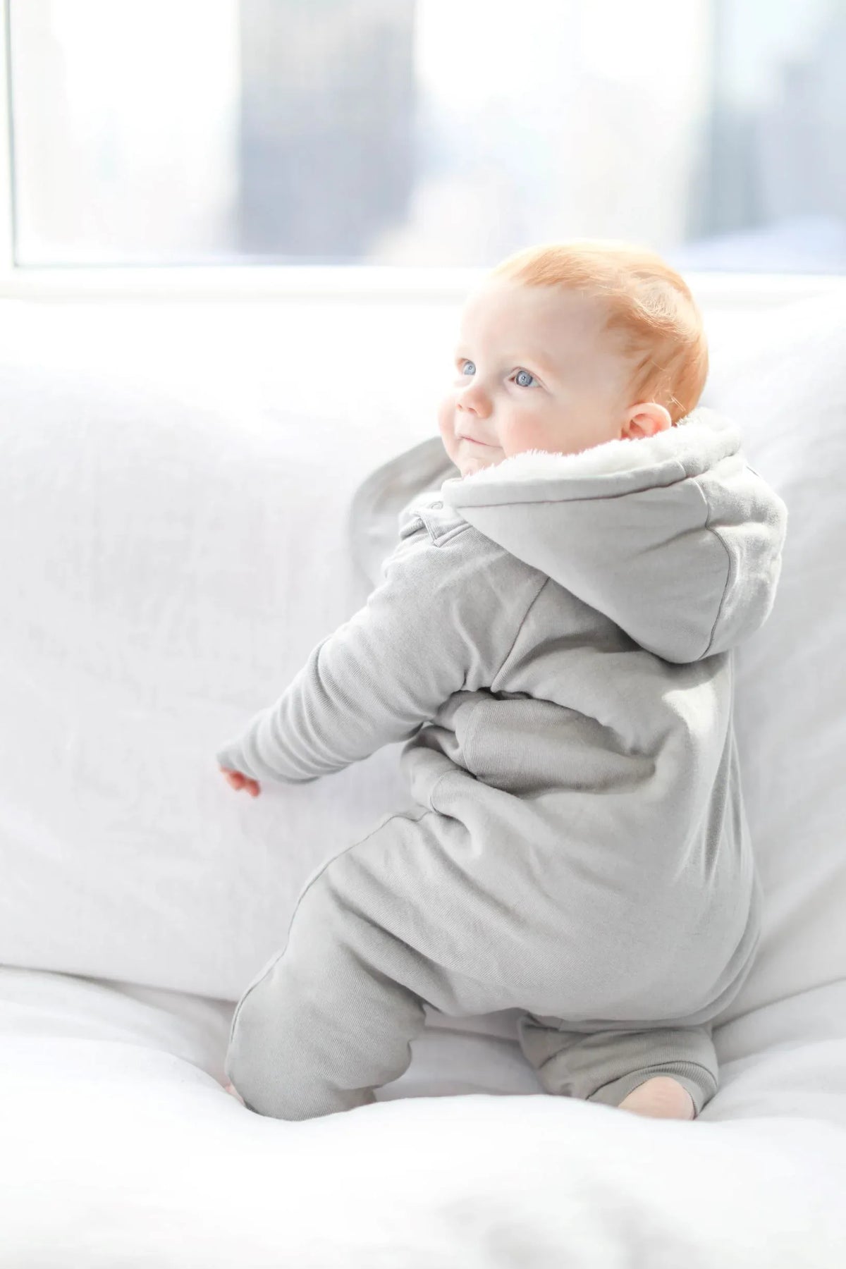 Baby smiling wearing gray jumpsuit with bib, feet covered, sitting against a plain background.