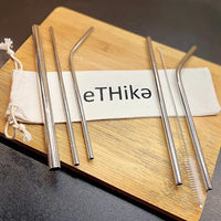Stainless steel straws arranged with cleaning brush and green cotton pouch on a wooden surface.