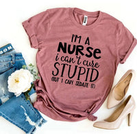 Flat lay of black T-shirt with white text reading 'I'm a Nurse I Can't Cure Stupid' on wooden background.