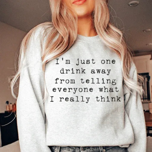 Front view of peach sweatshirt featuring bold 'I'm Just One Drink Away' text design on soft fleece fabric.