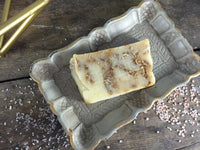 Oat and honey soap bar with textured surface on wooden soap dish with honey and oats nearby.