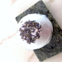 Hand holding a round lavender bath bomb with visible natural ingredients embedded.