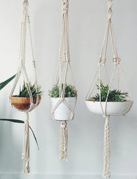 Hand-crafted macrame plant hanger in natural cotton rope holding a small potted plant against a white wall.