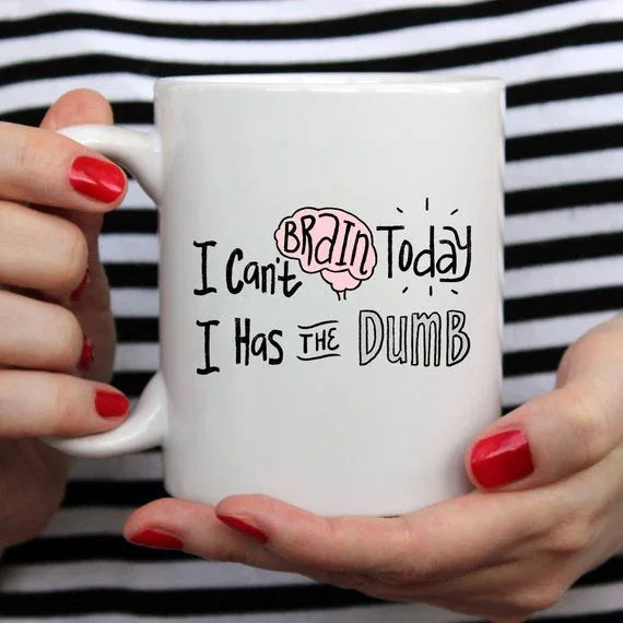 White coffee mug on a table with the phrase 'I Can't Brain Today, I Has The Dumb' visible.
