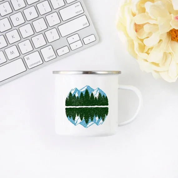White enamel mug next to camping gear, featuring mountain and forest illustration on both sides.