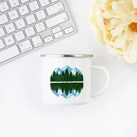 White enamel mug next to camping gear, featuring mountain and forest illustration on both sides.