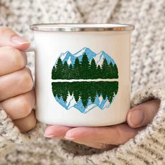 Enamel mug with scenic mountain print, set on wooden surface with natural light highlighting details.