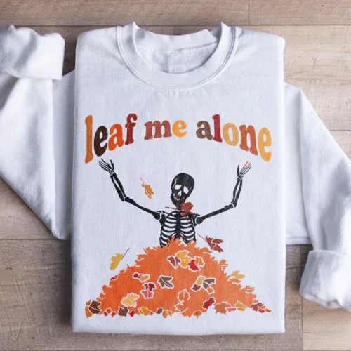 White sweatshirt with a stylized autumn leaf design in orange and yellow accompanied by Leaf Me Alone Fall text