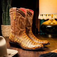 Man wearing mustard brown genuine caiman belly skin cowboy boots standing on wooden floor.